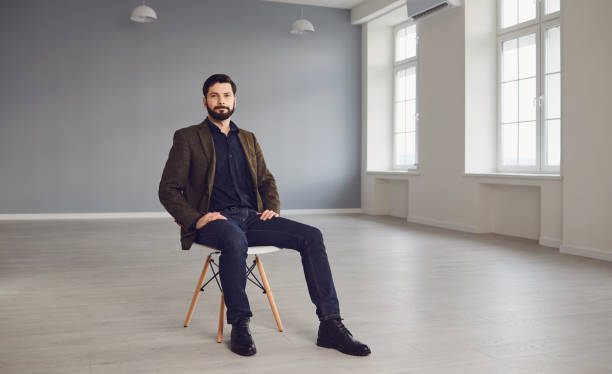 Elegant young man sitting in empty room Full length of confident bearded man in stylish jacket looking at camera while sitting on chair in empty modern studio sitting stock pictures, royalty-free photos & images