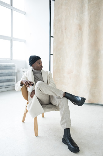 Fashion portrait of handsome young man wearing white coat, turtleneck and black beanie knit hat and boots sitting on chair in studio next to beige background.
