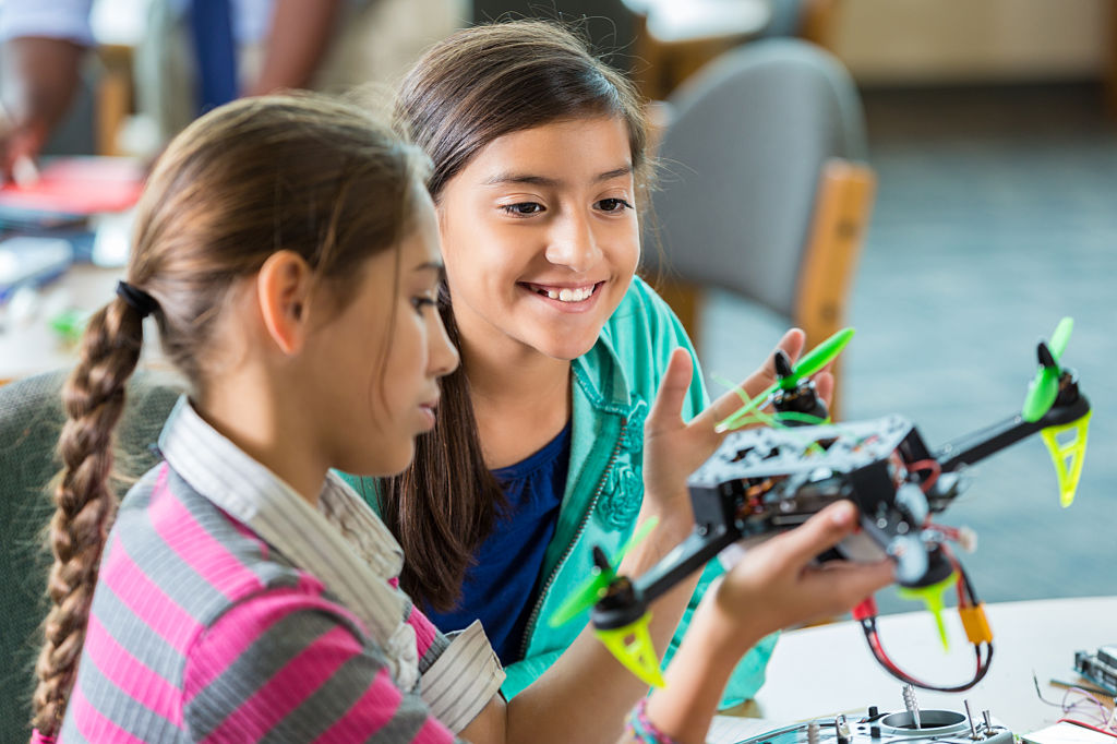 Elementary age Hispanic little girls are using drones during after school science club or program. Students are studying science, technology, engineering, and math in public elementary school library makerspace.