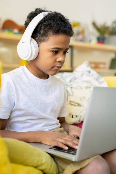 Elementary schoolboy of African ethnicity with headphones typing on laptop Serious elementary schoolboy of African ethnicity with headphones typing on laptop while listening to educational online course at home remote control stock pictures, royalty-free photos & images