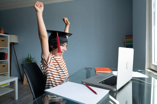 Elementary Schoolboy Using Laptop Computer And Wearing Mortarboard Photo of elementary schoolboy using laptop computer while wearing mortarboard. He is sittin on a desk next to window. shot indoor with a full frame mirrorless camera. online computer science degree stock pictures, royalty-free photos & images