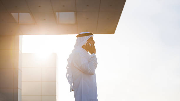 Emirati Businessman On A Rooftop In Evening Sunlight Traditionally dressed arab an is on a rooftop of a high building, talking on the phone and looking into the distance. Image is in letterbox format (16:9), contains copy space on both sides and has lot of intense vibrant sunlight. Made in Dubai, United Arab Emirates. middle eastern culture stock pictures, royalty-free photos & images