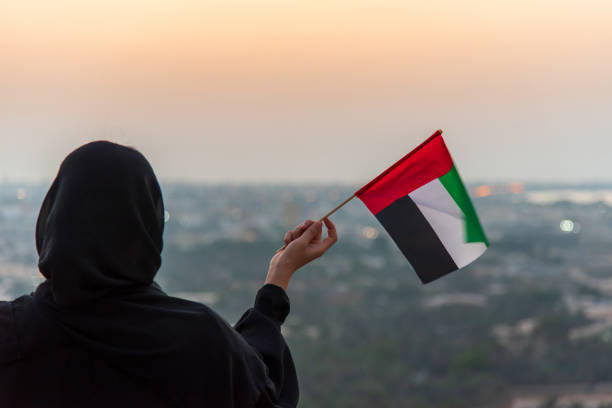 Emirati woman wearing hijab holding United Arab Emirates flag in the desert mountain at sunset