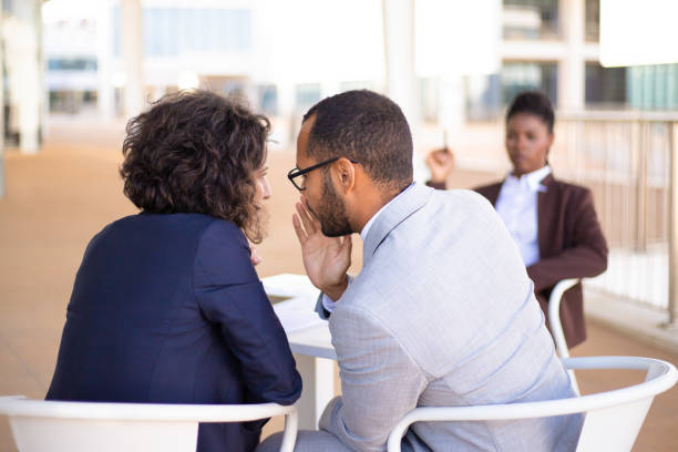 Employees gossiping about young female colleague Employees gossiping about young female colleague. Business man and woman whispering, African American employee sitting in background. Office rumors concept whispering stock pictures, royalty-free photos & images