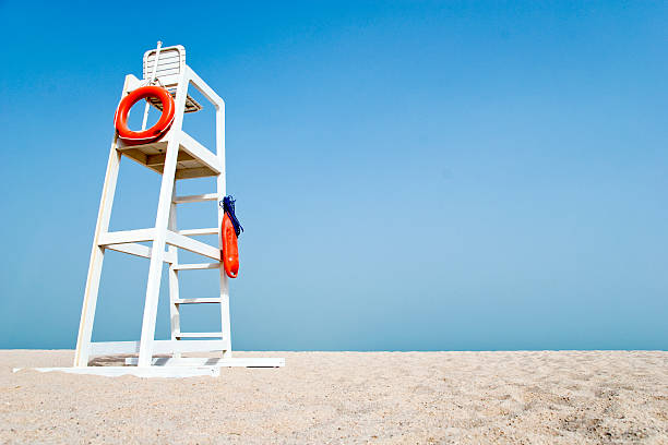 Empty Lifeguard Chair on the beach Empty white lifeguard chair on an empty beach with  orange life buoys hanging on the side.The chair stands against a clear blue sky in the sand. lifeguard stock pictures, royalty-free photos & images