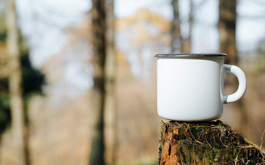 Empty metal hiking mug mock-up standing on tree stump in woods outdoors, copy space. Blank new enamelled cup for logo or branding, close-up.
