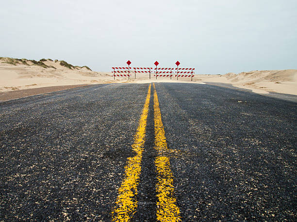 End of Road Last few feet of asphalt before red and white barriers indicate the end of the road.  Sand dunes line the road edges. dead end road stock pictures, royalty-free photos & images