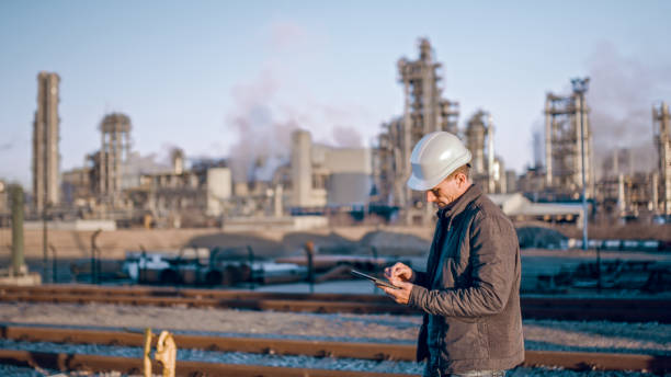 Engineer using tablet near oil refinery industry. Waist up shot of an engineer in a white hardhat using a tablet with an oil refinery visible in the background. oil industry stock pictures, royalty-free photos & images