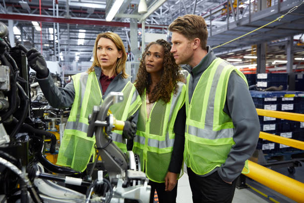 Engineers discussing over car part in factory Confident engineers discussing over car part in factory. Male and female colleagues are wearing reflective clothing. Professionals are working in car plant. car plant stock pictures, royalty-free photos & images