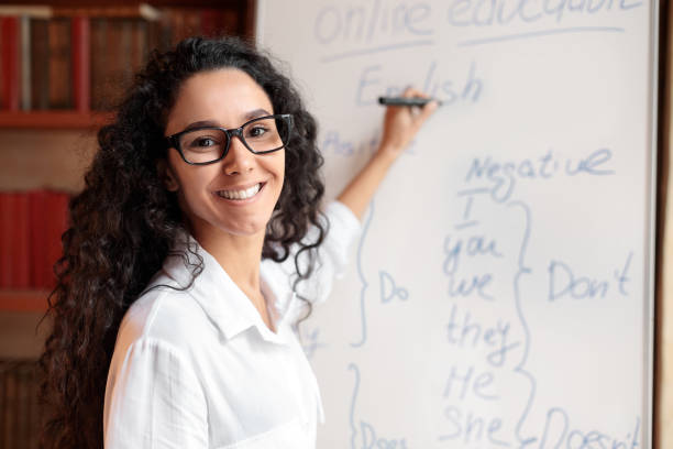 English teacher standing at board, explaining lesson to students Education And Learning Concept. Portrait of smiling female teacher standing at whiteboard, explaining grammar rules to students. Excited woman in glasses looking at camera, writing on the board showing stock pictures, royalty-free photos & images