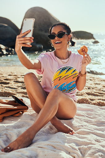 Shot of a woman taking a selfie while holding a donut on the beach