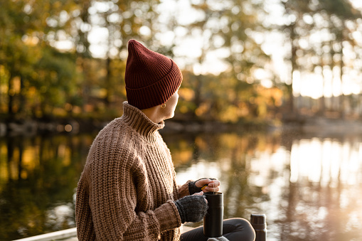 A woman preparing and drinking some fresh hot tea in a travel mug. She is sitting on a small jetty on the lake in autumn.