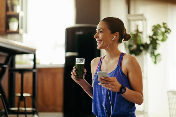 Enjoying in her healthy daily routine! Happy athletic woman listening music over earphones and drinking detox smoothie at home. drinking smoothie stock pictures, royalty-free photos & images