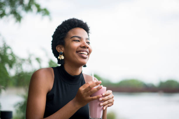 Enjoying The Summer Weather Beautiful Jamaican young woman wearing a black shirt is sitting outside by a lake while drinking a healthy strawberry smoothie. She is laughing with a toothy smile. drinking smoothie stock pictures, royalty-free photos & images