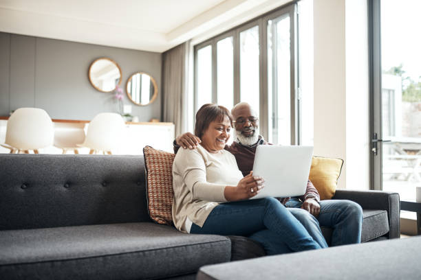 Entertained in the comfort of our own home Shot of a mature couple using a laptop while sitting together on the couch at home laptop couple stock pictures, royalty-free photos & images