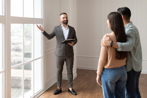 Look Here. Professional House Agent Seller In Suit Showing New Apartment To Young Couple, Pointing At Beautiful View From Floor to Ceiling French Windows To Embracing Future Homeowners. Purchase