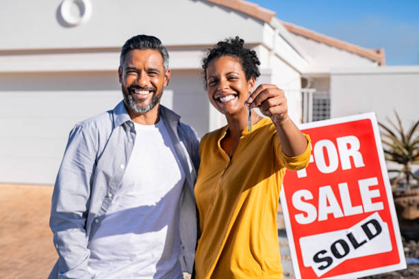 Ethnic couple holding new house keys Portrait of happy mixed race couple holding house keys standing near sold signboard. Middle eastern man embracing african woman while showing house key outside of their new home. Proud man and black girfriend moving home after buying their new apartment. selling stock pictures, royalty-free photos & images