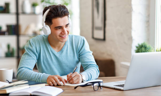 Ethnic student making notes during online lesson Happy ethnic guy in headphones smiling and writing in notebook while sitting at table and listening to teacher during online lecture at home online colleges stock pictures, royalty-free photos & images
