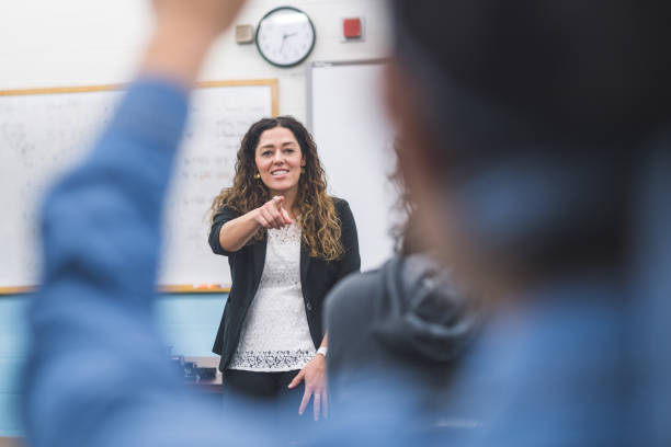 Ethnic teacher points to one of her students with a raised hand A teacher stands up front and calls on one of her students to respond to a question under discussion. Several hands are raised. high school teacher stock pictures, royalty-free photos & images