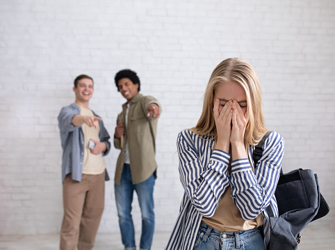 Bullying, negativity and peer problems. European and african american teens zoomers guys laugh at crying female with backpack in school or college, on white wall background, studio shot, empty space