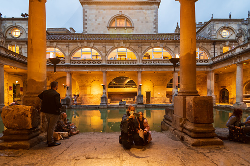 City of Bath, UK. 07-08-2021. Evening sightseeing of restored in Victorian times ancient Roman Baths in the light of wall sconces. Parent with child taking selfie on mobile phone.
