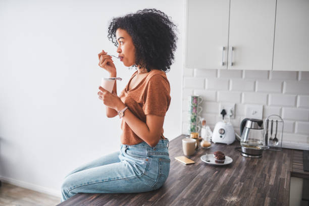 Every spoonful tastes like more Shot of a young woman having a tub of yoghurt in the kitchen at home breakfast photos stock pictures, royalty-free photos & images