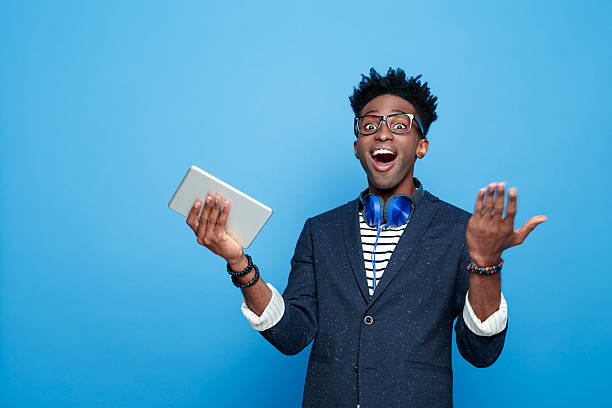 Excited afro american guy in fashionable outfit, holding digital tablet Studio portrait of surprised afro american young man wearing striped top, navy blue jacket, nerd glasses and headphone, staring at camera with mouth open, holding a digital tablet in hand. Studio portrait, blue background. good news stock pictures, royalty-free photos & images