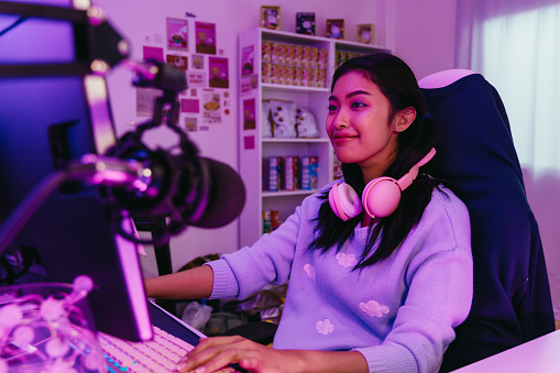 Excited and smiling gamer girl in cute headset with a mic playing an online video game and live streaming. Young Asian woman talking to players and audience on computer in neon led light room at home