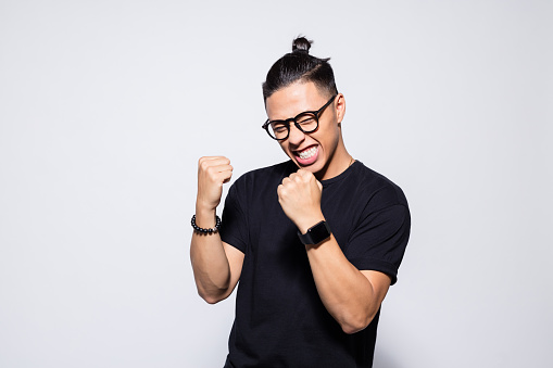 Portrait of happy asian young man wearing black t-shirt and eyeglasses, clenching fists. Studio shot, grey background.