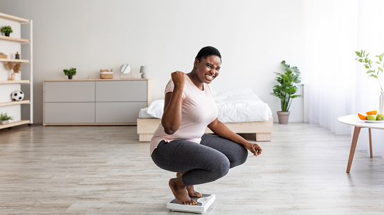 Excited curvy black woman sitting on scales at home, making YES gesture, happy with result of her slimming diet, panorama. Overjoyed plus size African American lady achieving her weight loss goal