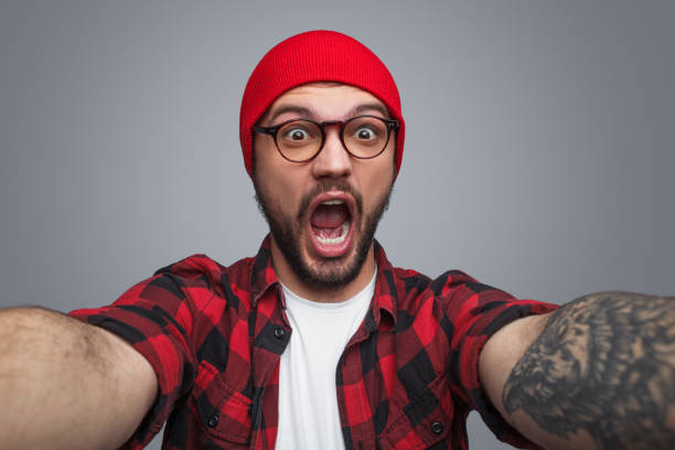Excited handsome man taking selfie in studio Amazed young unshaven male in glasses and red knitted beanie holding camera and making selfie looking at camera with open mouth over grey studio background mouth open stock pictures, royalty-free photos & images