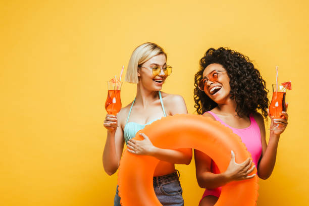 excited interracial women with swim ring and cocktail looking at each other on yellow - friends color background imagens e fotografias de stock
