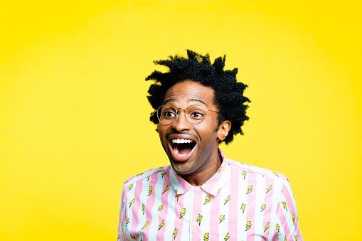 Summer portrait of happy afro american young man with dreadlocks wearing vintage shirt with lightning pattern and gold glasses, looking away with mouth open. Studio shot on yellow background.