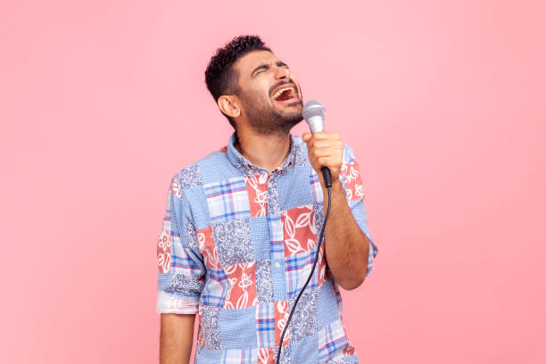 Excited positive man with beard in casual blue shirt singing songs, holding microphone, singer making performance, keeps eyes closed. stock photo