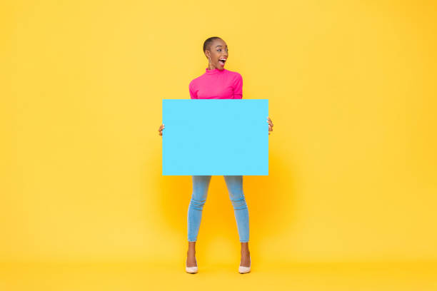 Excited pretty African American woman holding blue paper board with empty space for text on yellow isolated studio background stock photo