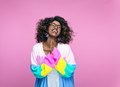 Cheerful african young woman wearing rainbow cardigan looking up and laughing. Studio portrait on pink background.