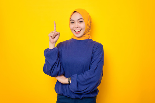 Excited young Asian Muslim woman dressed in casual sweater pointing finger up, having an idea or found solution isolated over yellow background