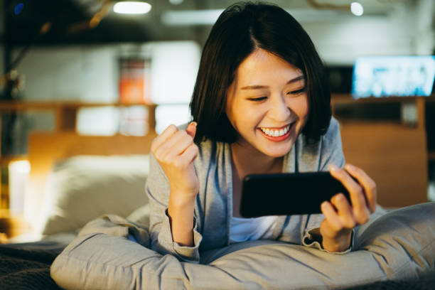 興奮した若いアジアの女性は、自宅で夕方にスマートフォンでモバイルゲームをプレイし、寝室のベッドに横たわっている - ゲーム ストックフォトと画像