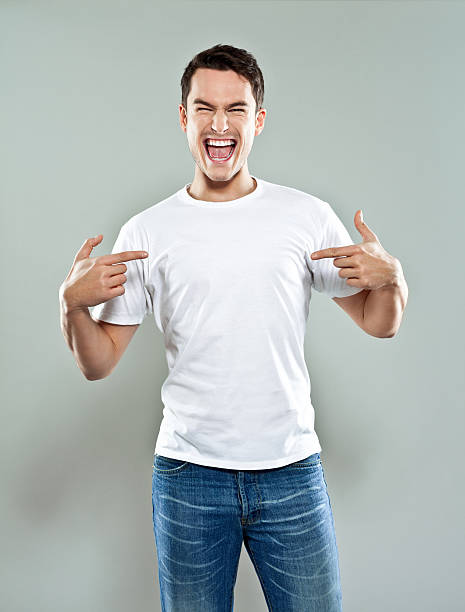 Excited young man Portrait of happy young man wearing white t-shirt, pointing with fingers at his torso and laughing at camera. Studio shot, grey background. one man only stock pictures, royalty-free photos & images