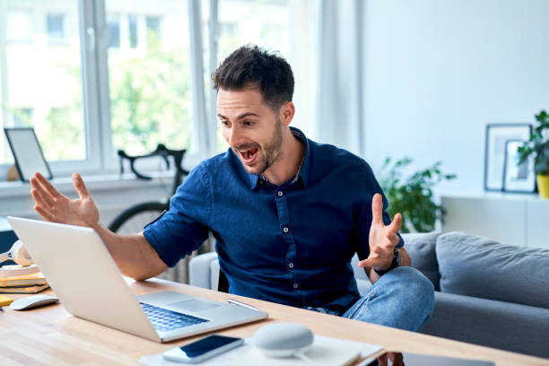 Excited young man shouting at laptop while working at home stock photo