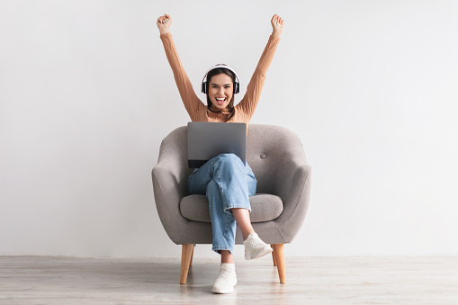 Excited young woman in headphones sitting in armchair, celebrating online win, great deal or business success. Millennial lady enjoying big sale in web store, feeling victorious