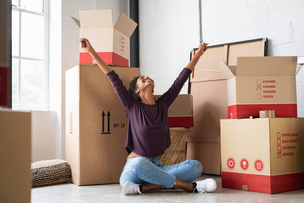 Excited young woman in new house Happy young woman sitting in new apartment and raising arms in joy after moving in. Joyful and excited african girl moving to new home. Black woman sitting on floor in her house. relocation stock pictures, royalty-free photos & images