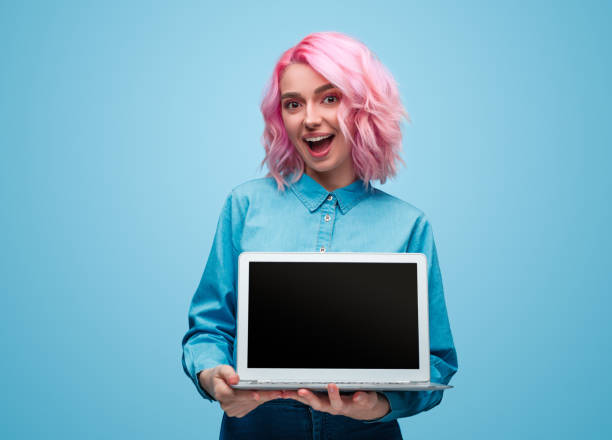 Excited young woman showing laptop Amazed young lady with pink hair looking at camera and demonstrating modern laptop with blank screen while standing on blue background pink hair stock pictures, royalty-free photos & images