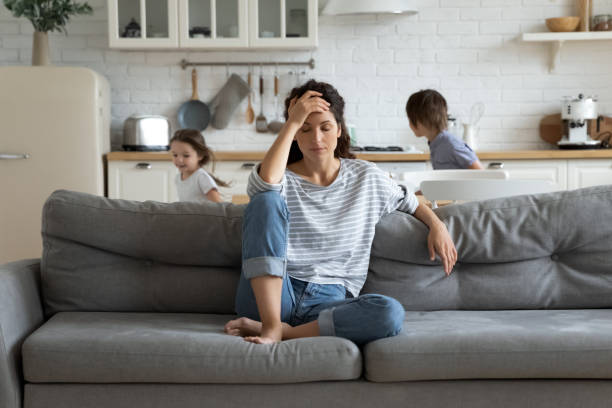 Exhausted mother sitting on couch while kids running at home Young tired single mother suffers from headache closed eyes touch forehead sitting on couch while her daughter and son running around her and shouting, female babysitter feels exhausted by noisy kids headache stock pictures, royalty-free photos & images