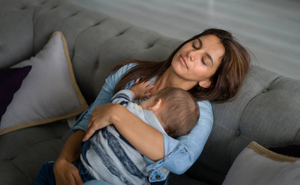 Exhausted mother sleeping in the sofa whole holding her baby stock photo