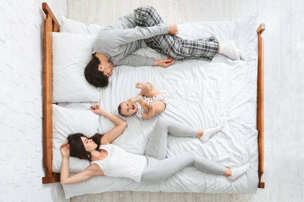 Exhausted parents sleeping on sides of bed, active baby playing in middle Joint sleep with kids. Exhausted parents sleeping on sides of bed, active baby playing in middle, above view man sleeping in bed top view stock pictures, royalty-free photos & images