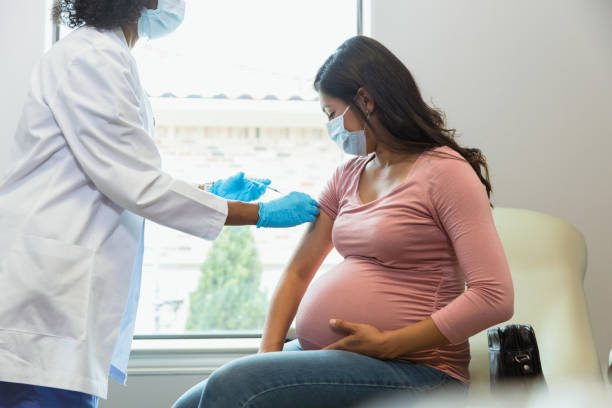 Expectant mother watches as tech gives her vaccine Wearing protective masks because of COVID-19, a mid adult pregnant woman watches as the female technician administers a booster shot. obstetrician photos stock pictures, royalty-free photos & images