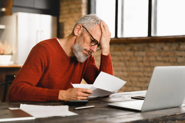 Expensive charges on domestic bills. Loan, debt, bunkruptcy concept. Sad depressed caucasian businessman holding documents, having problems with dismissal at home office stock photo