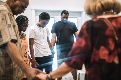 A large multi-generational group of family relatives holds hands and prays together in a living room, having not seen each other for a long time, due to normal circumstances or perhaps COVID-19 or Coronavirus.  Faith and connection are powerful for hope.