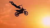 SILHOUETTE: Pro motocross rider riding fmx motorbike, jumping big air kicker performing extreme stunt. Professional biker jumps no hander superman trick over golden sunset sky above trees